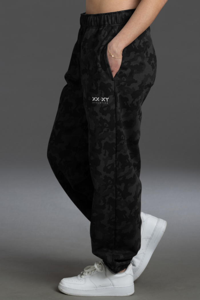 Black camouflage pants with a brand logo worn by a person on a gray background || Black Camo