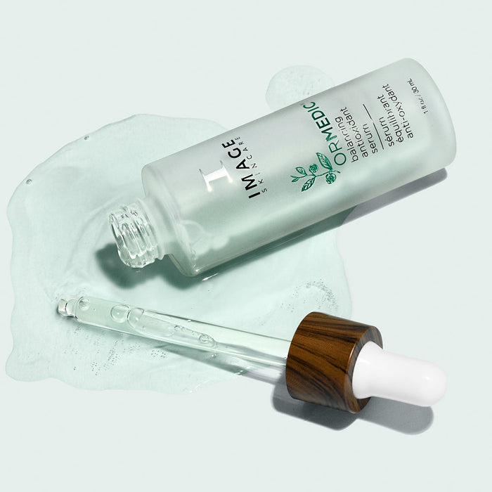 Clear glass dropper bottle labeled ORMADIC balancing antioxidant serum | IMAGE Skincare laying on a pale surface with serum droplets and a wooden-capped dropper nearby, showcasing skincare product and wellness supplement.