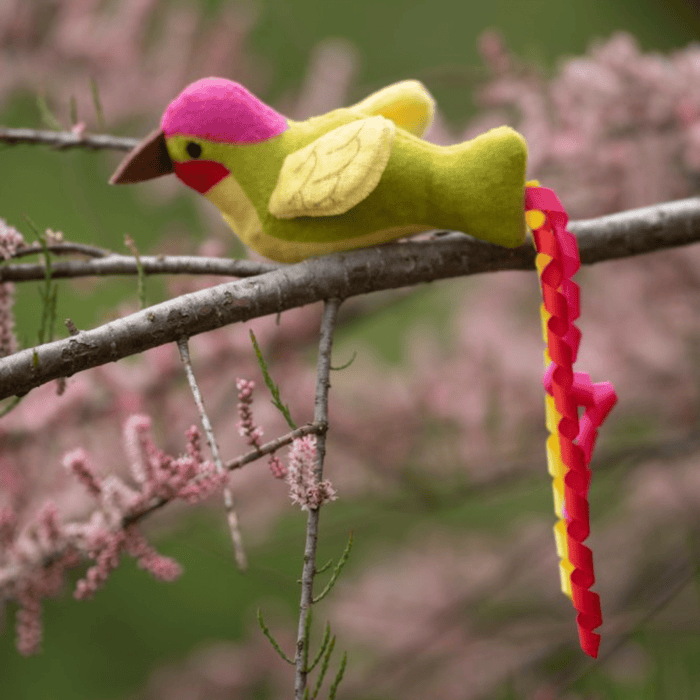 Green, pink and yellow Hummingbird cat toy with a three curls of ribbon as tail in a tree with pink blossoms.