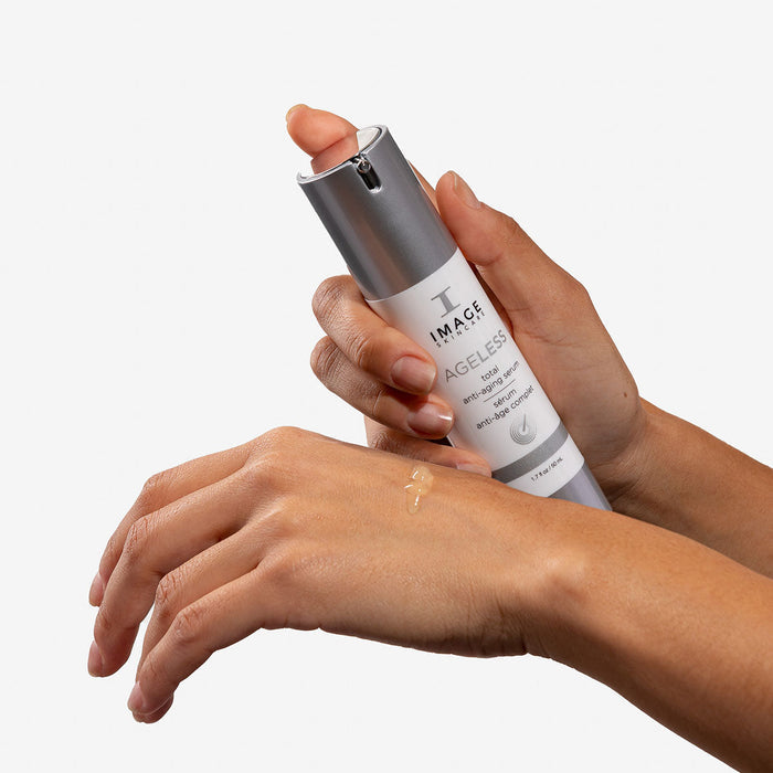 Hand holding a bottle of AGELESS total anti-aging serum | IMAGE Skincare with a small amount of serum applied on the back of the other hand, showcasing skincare application.
