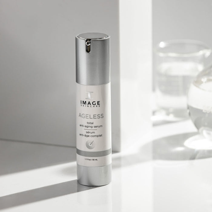 Bottle of AGELESS total anti-aging serum | IMAGE Skincare on a white surface with soft natural light and glassware in the background, highlighting skincare and wellness.