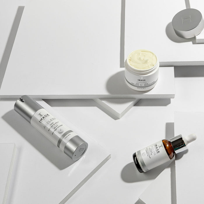 Three IMAGE Skincare products on white geometric surfaces, featuring AGELESS total anti-aging serum | IMAGE Skincare with a dropper bottle, a cream jar, and a silver pump bottle for skincare and wellness.