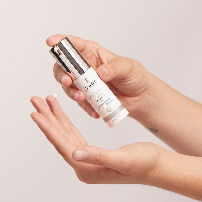 Hand holding a bottle of AGELESS total eye lift crème | IMAGE Skincare with a small amount of cream dispensed onto the fingertip, showcasing skincare application.