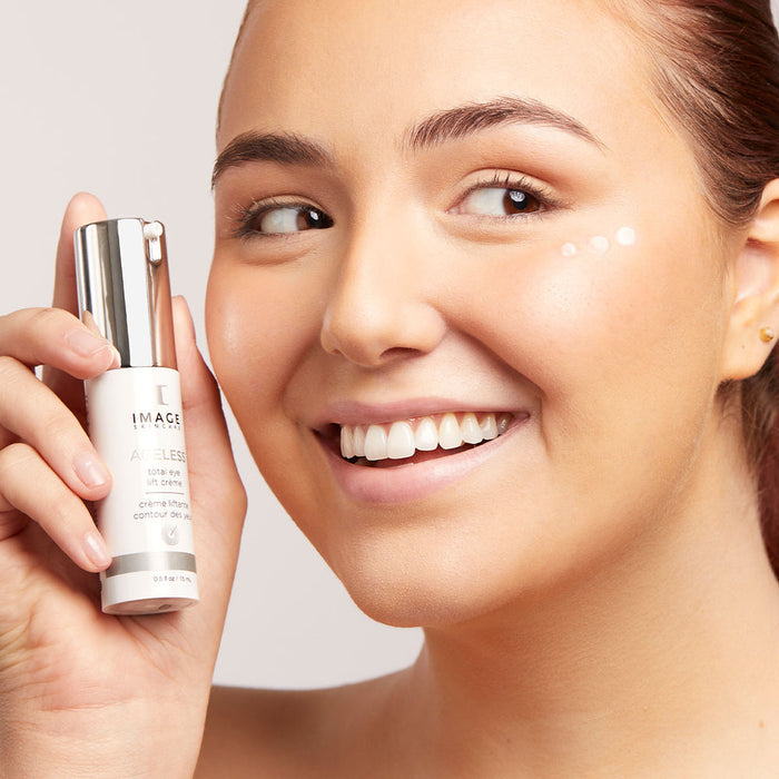 Smiling woman holding a bottle of AGELESS total eye lift crème | IMAGE Skincare with small dots of the crème applied under her eye in a bright skincare setting.