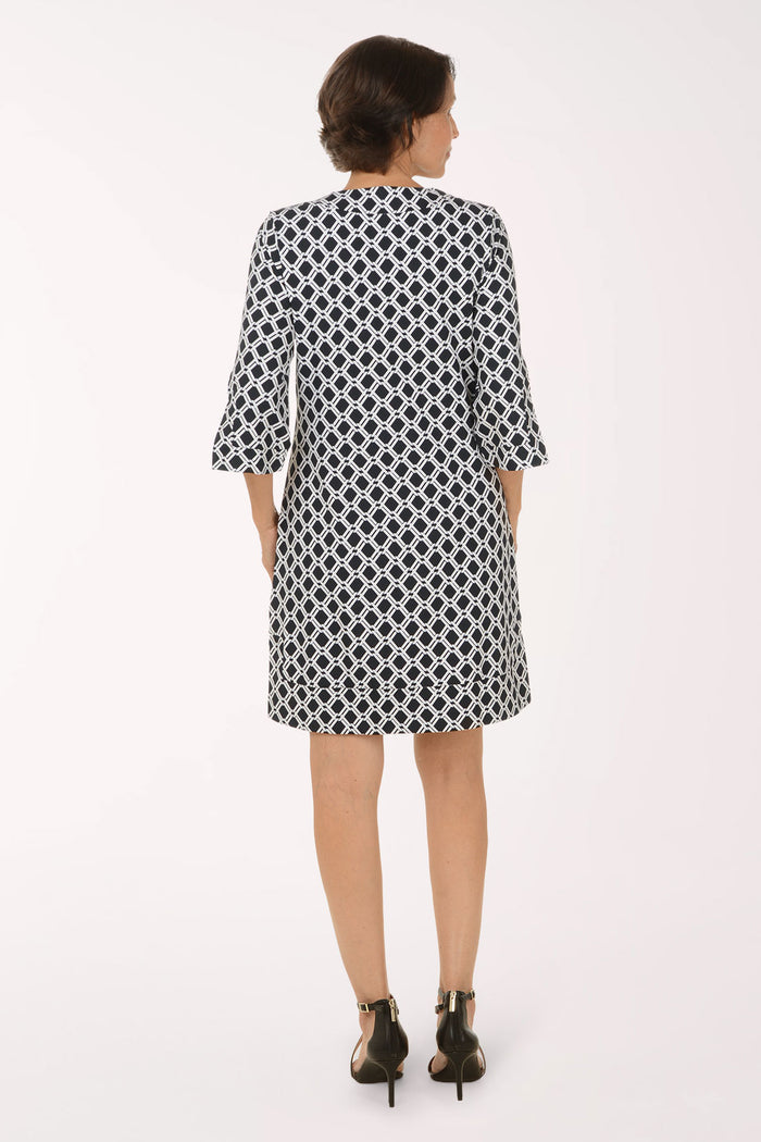 A woman modeling a short black and white geometric patterned dress with elbow-length sleeves and a split neckline, paired with black strappy high-heeled sandals, against a plain white studio background.