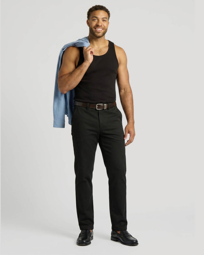 A smiling man wearing a black Ribbed Curved Hem Tank 3-Pack top paired with black pants, holding a light blue jacket over his shoulder, showcasing casual men's basics style.