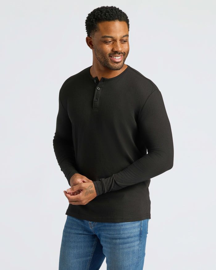 Man wearing Black Long Sleeve Henley, adjusting sleeve, with a slight smile.