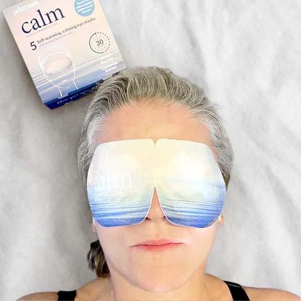 Woman lying down wearing a Calm Self Warming Pressure Point Eye Mask - Single | Popmask with a soothing blue and white gradient design, next to its calming packaging.