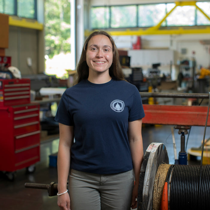 Woman wearing a navy blue t-shirt with a white WHOI Classic Seal Logo on the front left chest in an industrial work area.