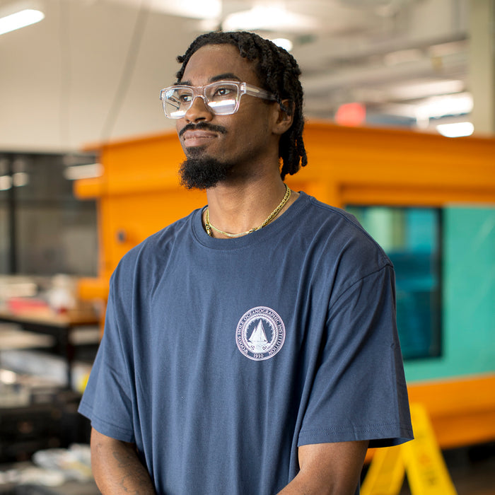 Man wearing a navy blue t-shirt with a white WHOI Classic Seal Logo on the front left chest in a lab.