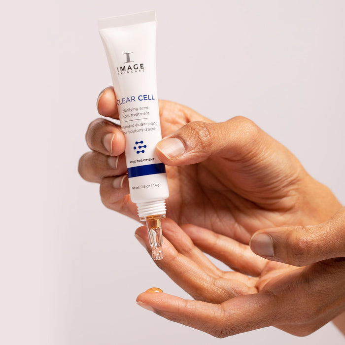 Hand holding a tube of CLEAR CELL clarifying acne spot treatment | IMAGE Skincare with a drop of clear gel dispensed onto the fingertip against a neutral background.