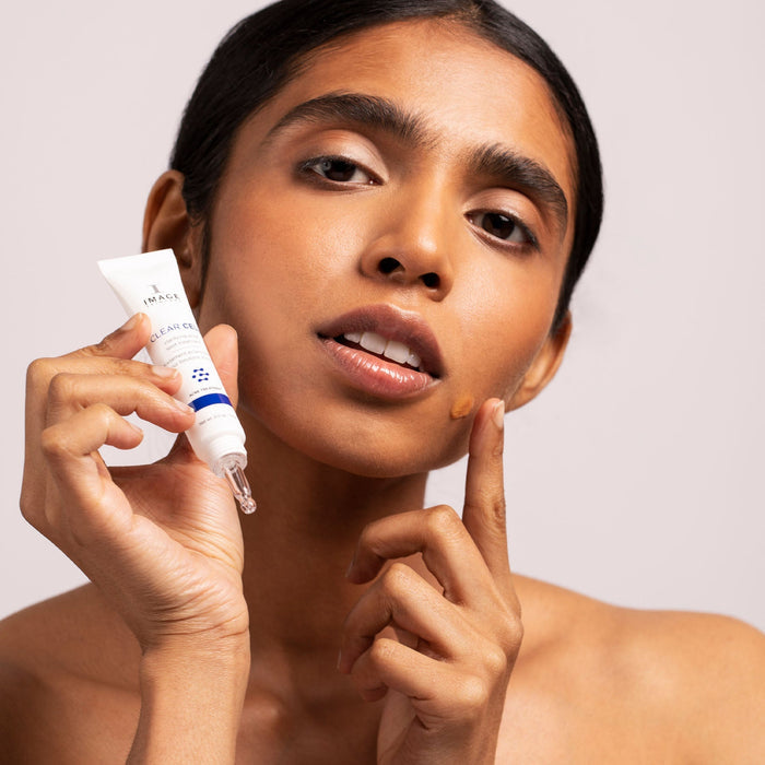 Woman with clear skin applying CLEAR CELL clarifying acne spot treatment | IMAGE Skincare on her cheek, demonstrating targeted skincare for acne treatment.