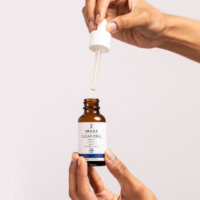 Hands holding a dropper and a brown glass bottle of CLEAR CELL Restoring Serum | IMAGE Skincare against a neutral background, showing skincare product application.