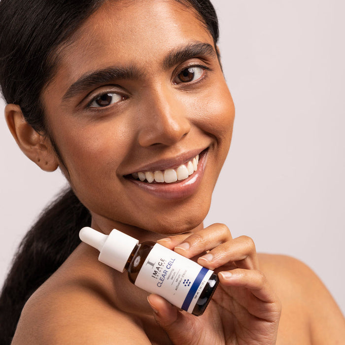 Smiling woman with clear skin holding a bottle of CLEAR CELL Restoring Serum | IMAGE Skincare, showcasing the skincare product for healthy, glowing skin.