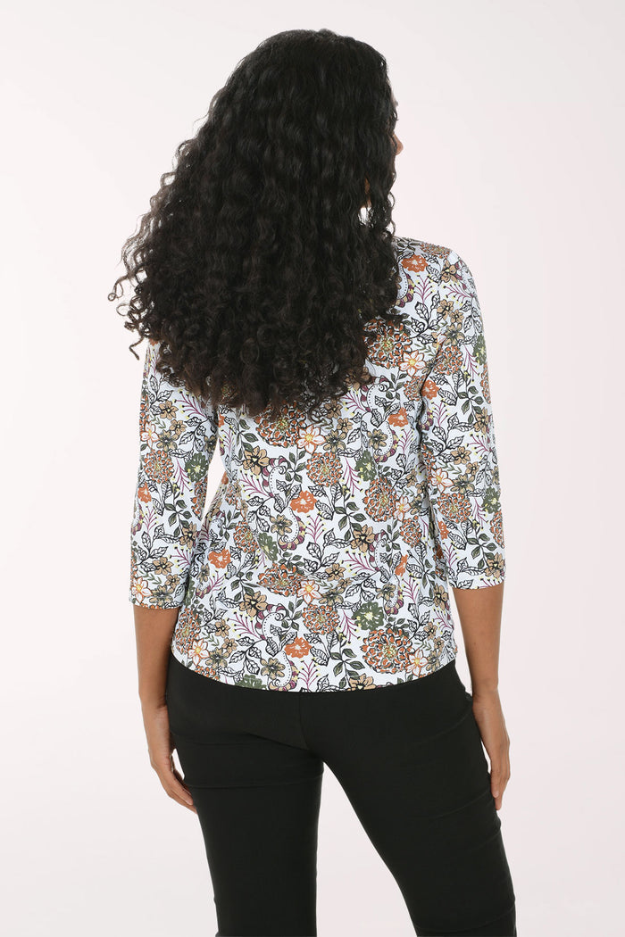 A woman with long curly black hair wearing a white three-quarter sleeve top with a colorful floral and paisley pattern, paired with black pants, standing against a plain white background.