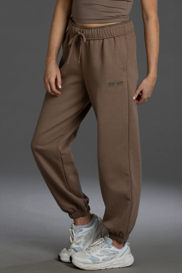 A person wearing brown jogger sweatpants with an elastic drawstring waist and elastic cuffs, paired with white sneakers, shown from the waist down against a dark gray studio background. || Mocha