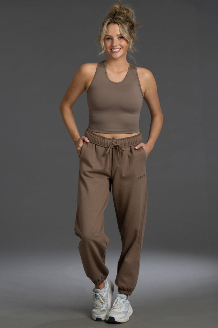 A woman modeling a matching taupe-brown activewear set consisting of a ribbed tank top and jogger sweatpants, paired with white sneakers, against a solid gray studio background. || Mocha