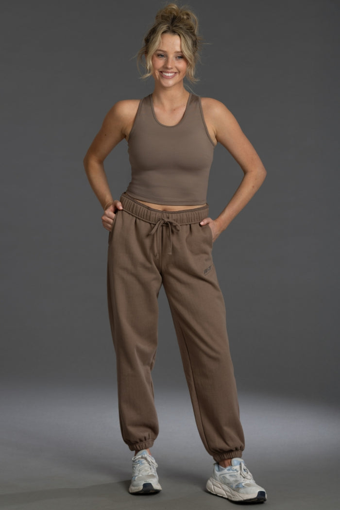A woman modeling a matching brown activewear set consisting of a cropped tank top and jogger sweatpants, paired with white sneakers, against a solid gray studio background. || Mocha