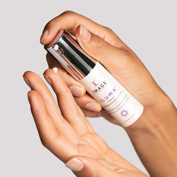 Person applying a small amount of ILUMA intense brightening eye crème | IMAGE Skincare from a sleek white and silver bottle onto their fingertip, highlighting skincare routine.