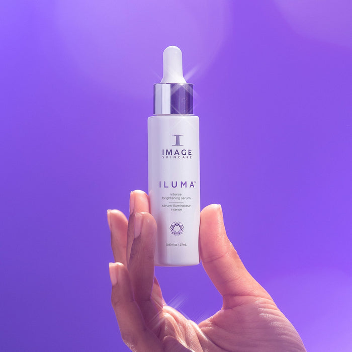 Hand holding a white dropper bottle of ILUMA® intense brightening serum | IMAGE Skincare against a soft purple background with light reflections, highlighting skincare and wellness.
