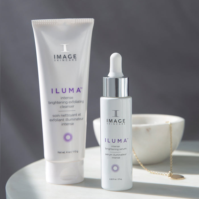 A white tube and a white dropper bottle of ILUMA® intense brightening serum | IMAGE Skincare displayed on a white surface with soft natural lighting and a blurred white bowl with a gold necklace in the background.