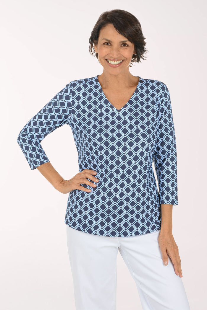 A woman modeling a blue and white geometric print long-sleeve top with a V-neck, paired with white pants, against a plain light background.