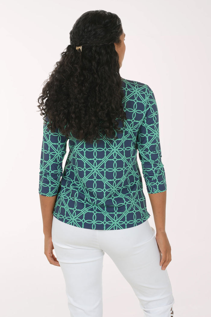A woman modeling a three-quarter sleeve V-neck top with a teal and navy geometric interlocking circle pattern, paired with white pants, against a plain white studio background.