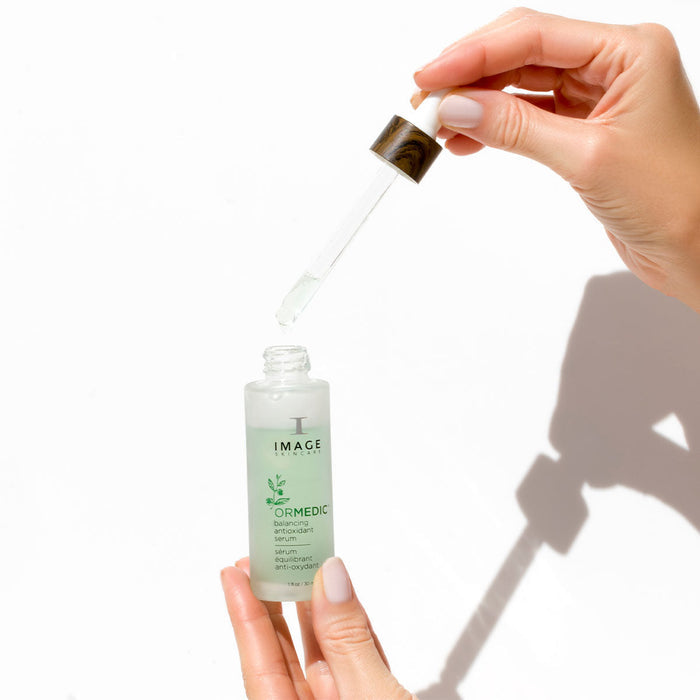 Hands holding a dropper and a bottle of ORMEDIC balancing antioxidant serum | IMAGE Skincare with clear liquid, highlighting skincare and wellness care rituals.