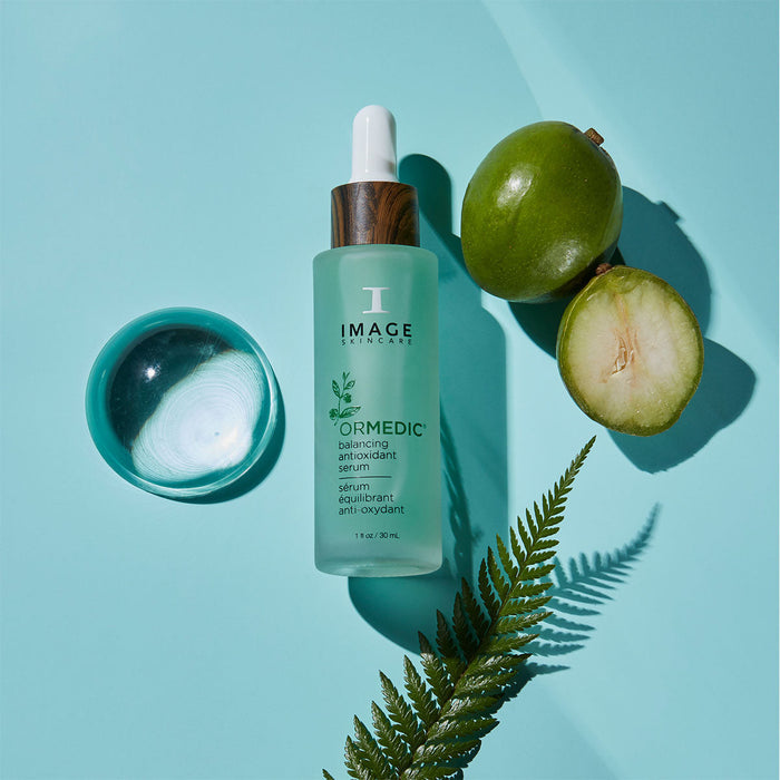ORMEDIC balancing antioxidant serum | IMAGE Skincare bottle with wooden cap next to a small bowl of clear liquid, green fruit halves, and a fern leaf on a light blue background.