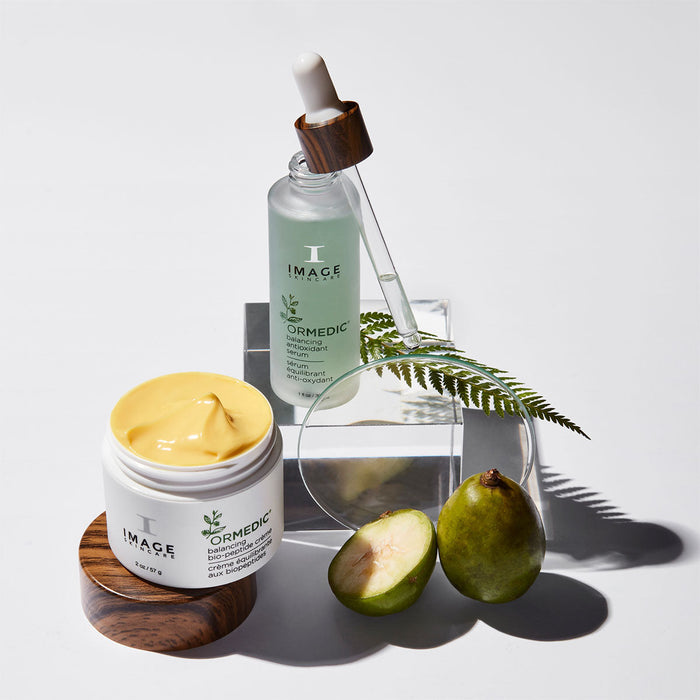 ORMEDIC balancing antioxidant serum | IMAGE Skincare bottle with dropper, paired with a cream jar, fresh green fruit, and a fern leaf on a white background, showcasing natural skincare ingredients.