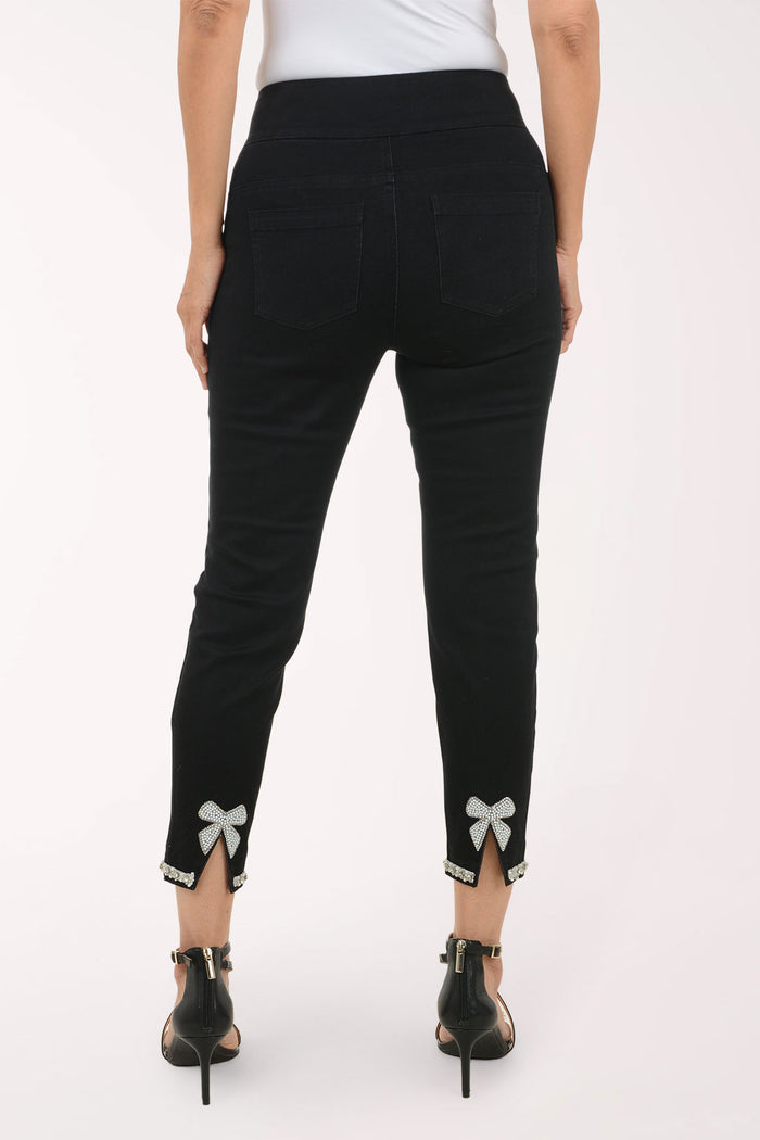 Rear view of a model wearing black cropped pants with silver bow details and side slits at the hem, paired with black high-heeled sandals against a plain white background.