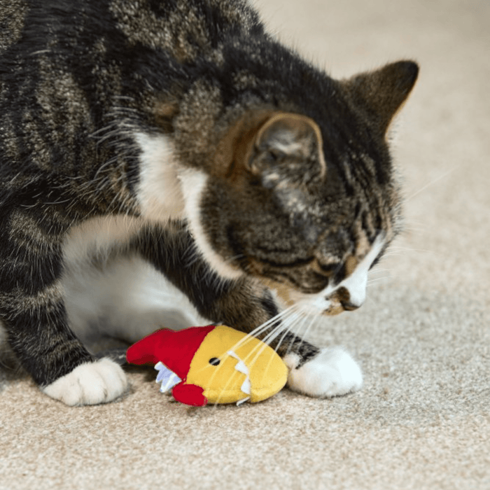 Brown tabby cat with white nose and paws playing with yellow and red Piranha cat toy with white felt teeth on carpet.