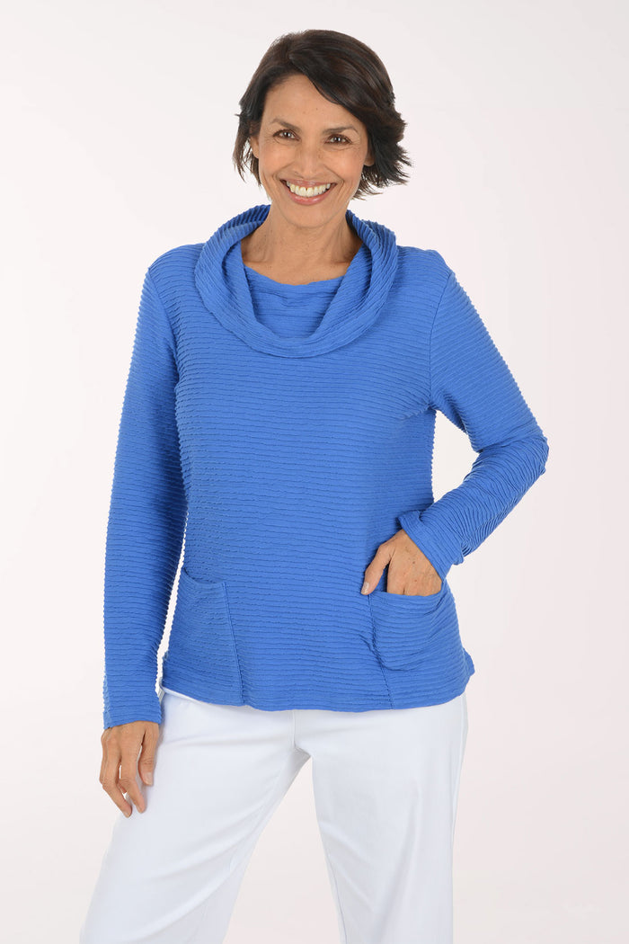 A smiling woman wearing a bright blue ribbed cowl-neck long-sleeve top with front patch pockets, paired with white pants, against a plain light background.