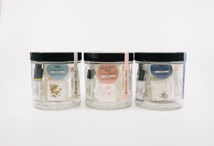Three clear jars with black lids, containing small vials, dried herbs, and labeled items, showcasing the Ritual Kit | Essence One for skincare and wellness.