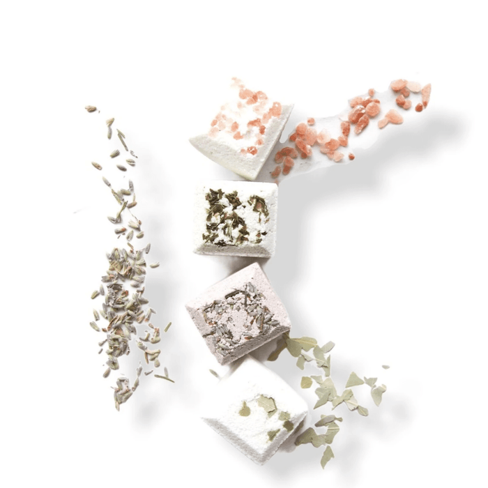 Four Shower Steamer | Essence One cubes arranged vertically with natural ingredients like dried lavender, pink Himalayan salt, herbs, and eucalyptus scattered around them on a white background.