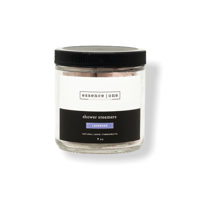 Clear glass jar with black lid containing lavender scented Shower Steamer | Essence One, labeled as natural, safe, and therapeutic, for body care and wellness.