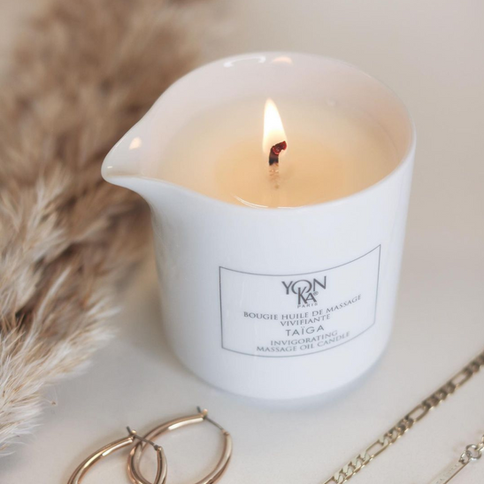 White ceramic candle burning with soft flame next to gold jewelry, labeled Taiga Massage Candle | Yon-Ka Paris for skincare and body care wellness.