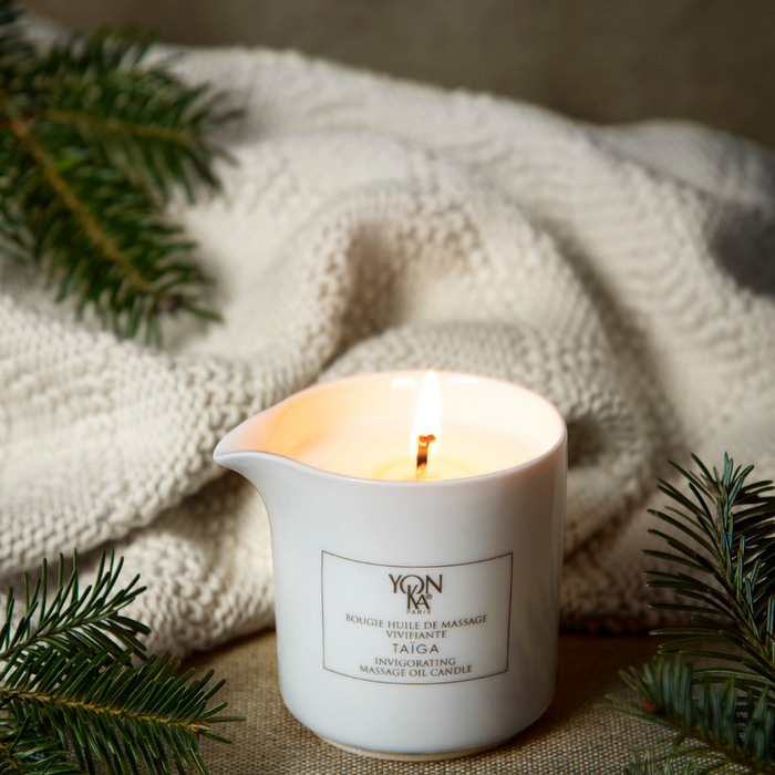 Lit Taiga Massage Candle | Yon-Ka Paris in a white ceramic container surrounded by pine branches and a cozy knitted blanket, evoking relaxation and skincare wellness.