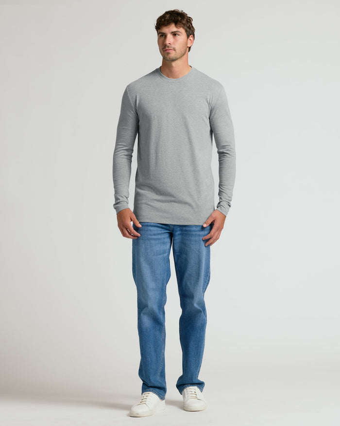 A man modeling a light gray long-sleeve crewneck t-shirt and blue jeans against a plain light gray studio background.