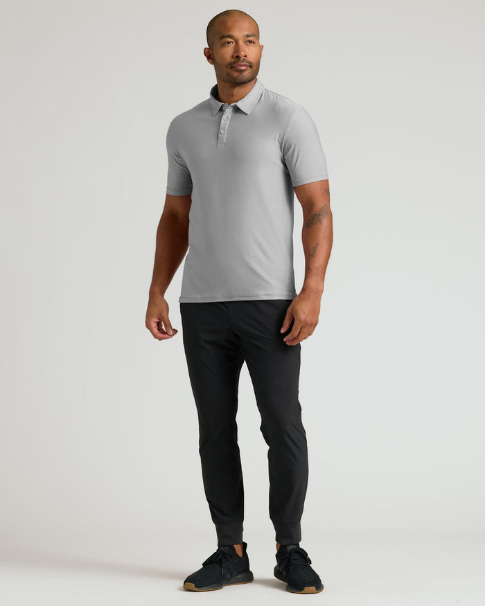 Man wearing a Light Heather Gray Active Polo 2.0 with short sleeves, paired with black pants and black sneakers, standing against a plain light background.