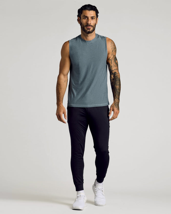 A muscular man wearing a heather navy Sleeveless Active Muscle Tee 3-Pack paired with black athletic pants and white sneakers, standing against a neutral background.